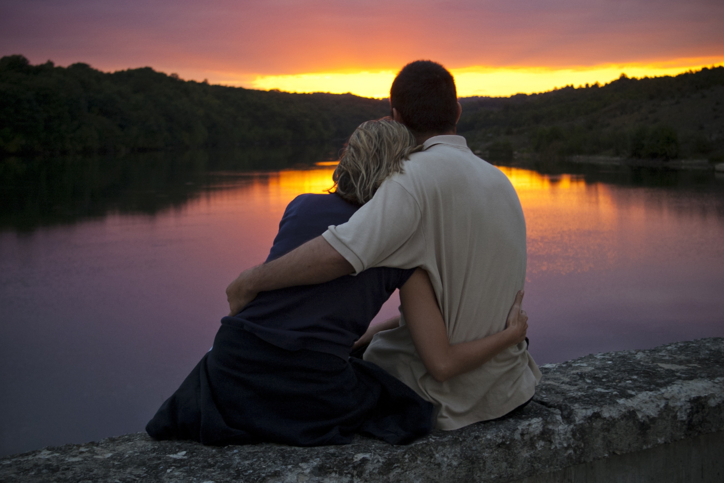 Девушка и парень обнявшись сидят на берегу реки и любуются красками вечерней зари