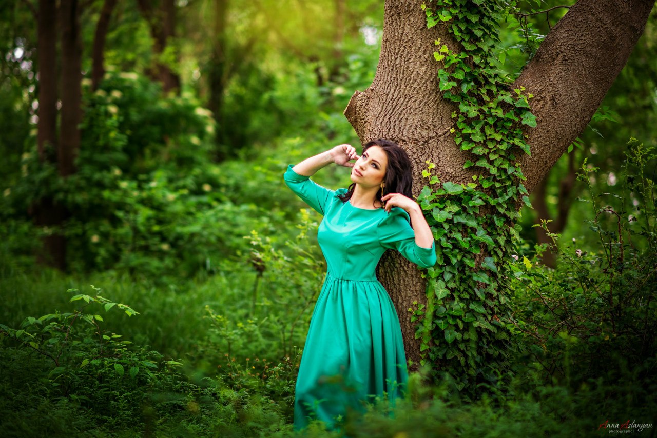 Девушка с длинными темными волосами стоит в закрытом бирюзовом платье, подняв руки к плечам. Она опирается на ствол дерева, которое растет в лесу.