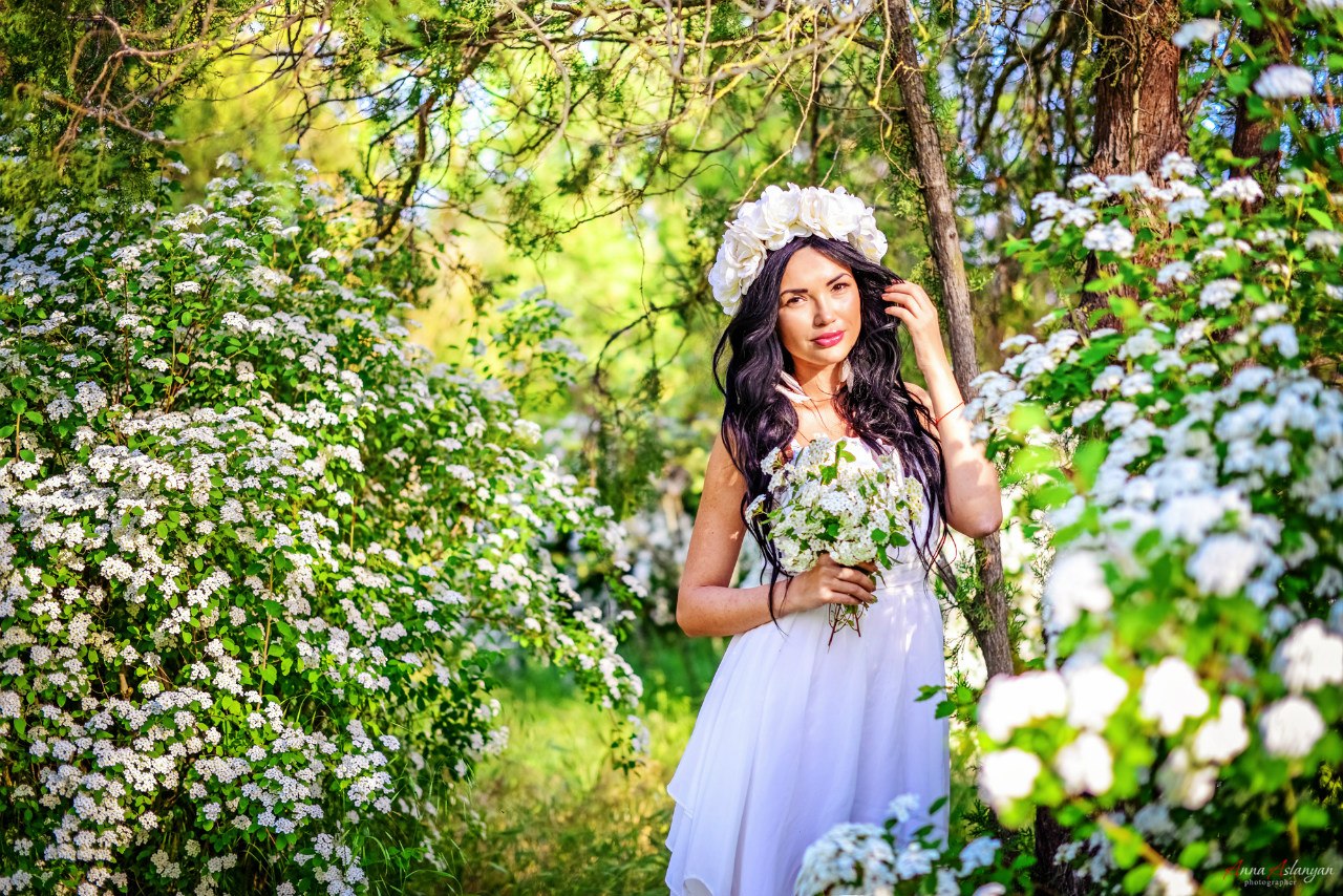Девушка брюнетка с длинными волосами в венке из белых цветов и в белом платье держит в руке букет из белых цветов и стоит среди цветущих кустов в лесу