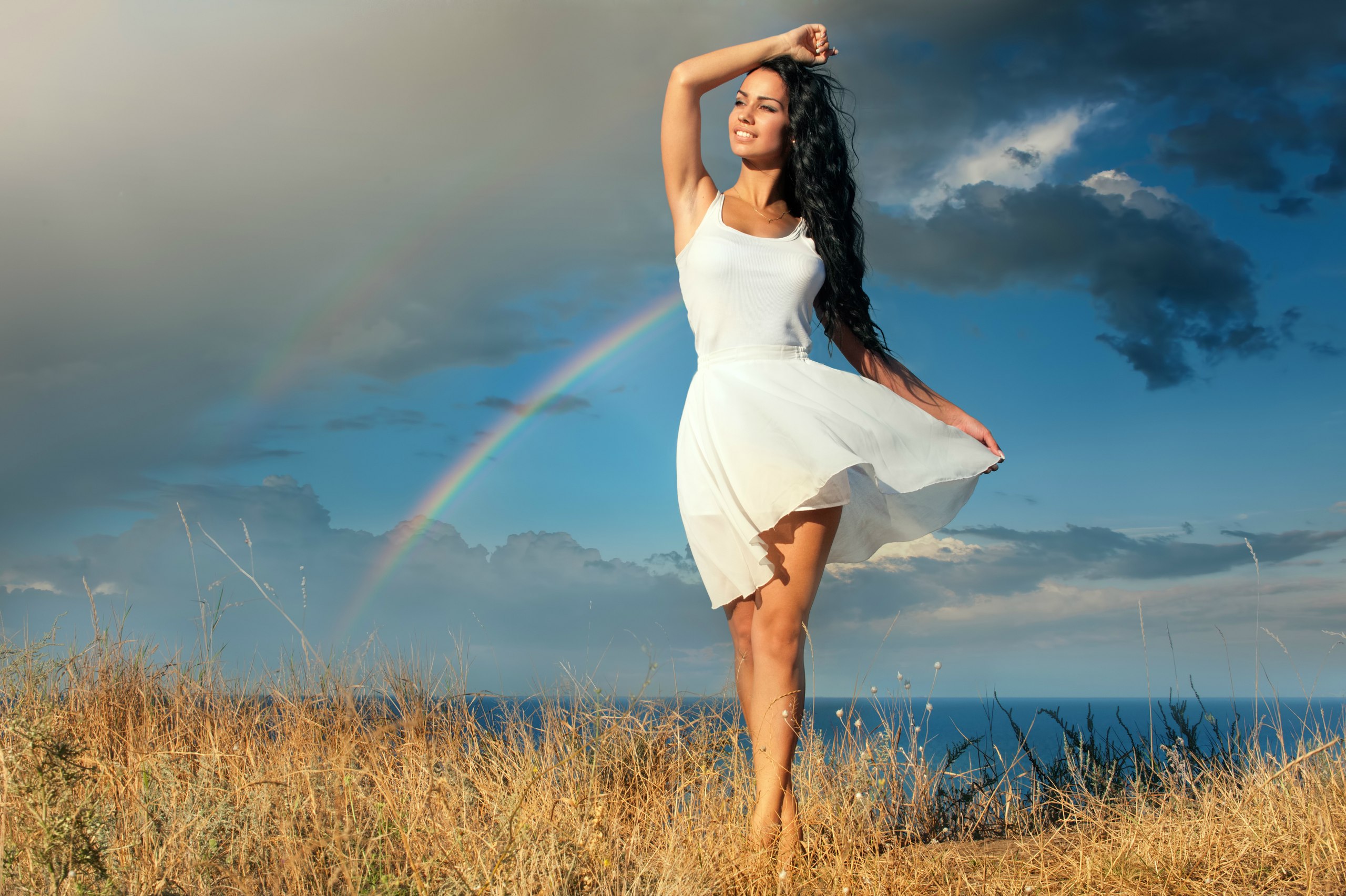 Девушка в белом платье стоит в поле на фоне неба в облаках и радуги