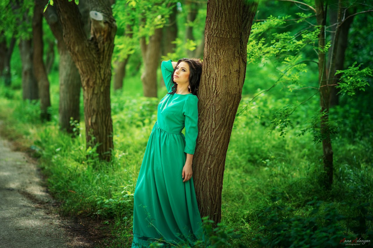 Девушка в бирюзовом платье стоит опираясь на ствол дерева в лесу, рядом с лесной дорожкой. Она задумчиво смотрит вверх. Фотограф Анна Асланян