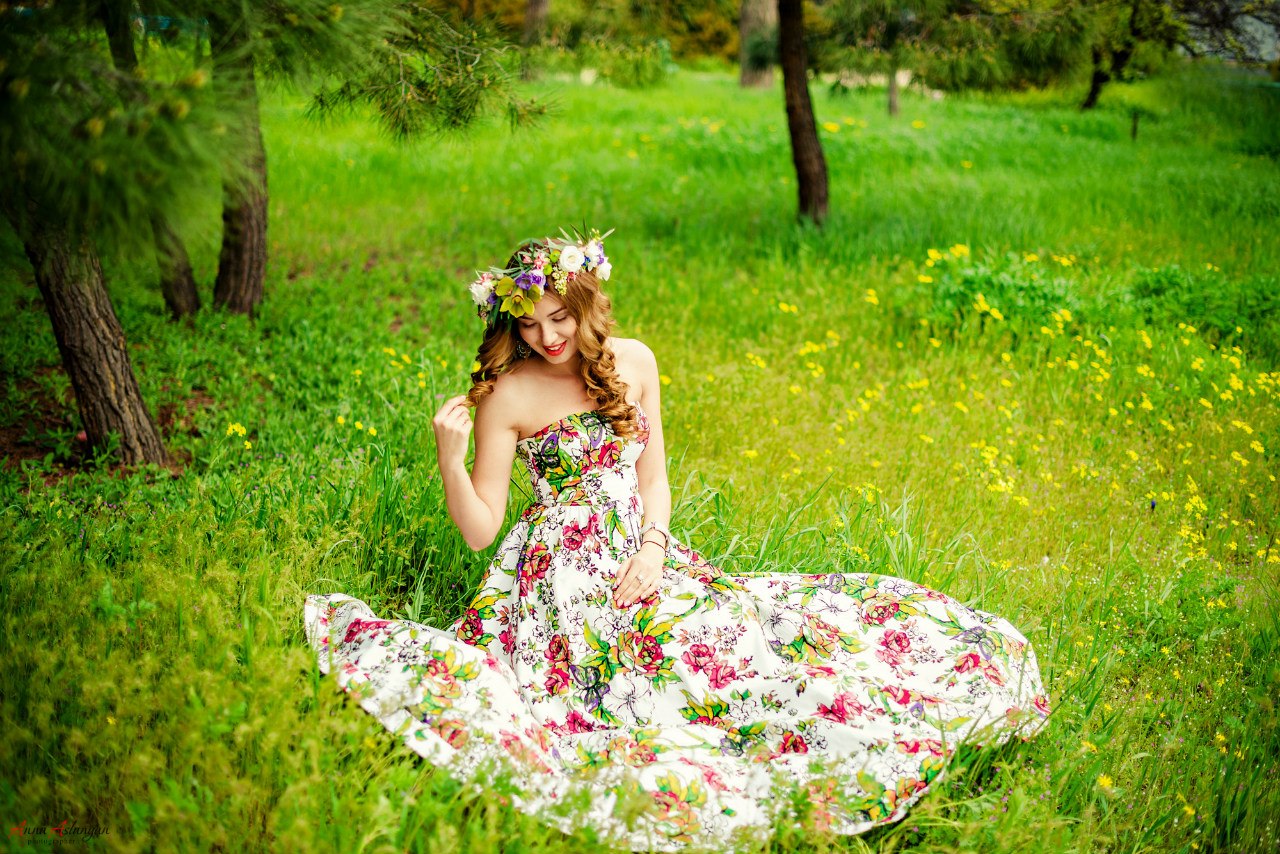 Девушка в венке из разноцветных цветов и в летнем платье с яркими цветами сидит на зеленой лужайке. Фотограф Анна Асланян