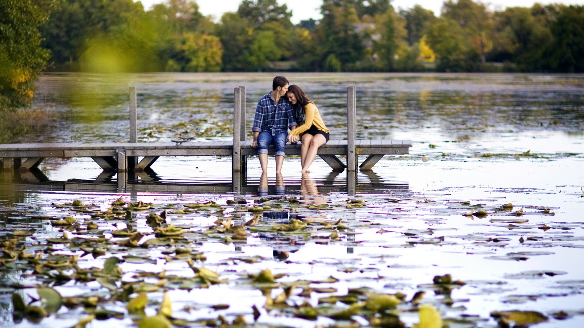 Парень и девушка обнявшись и опустив ноги в воду сидят на пирсе у озера
