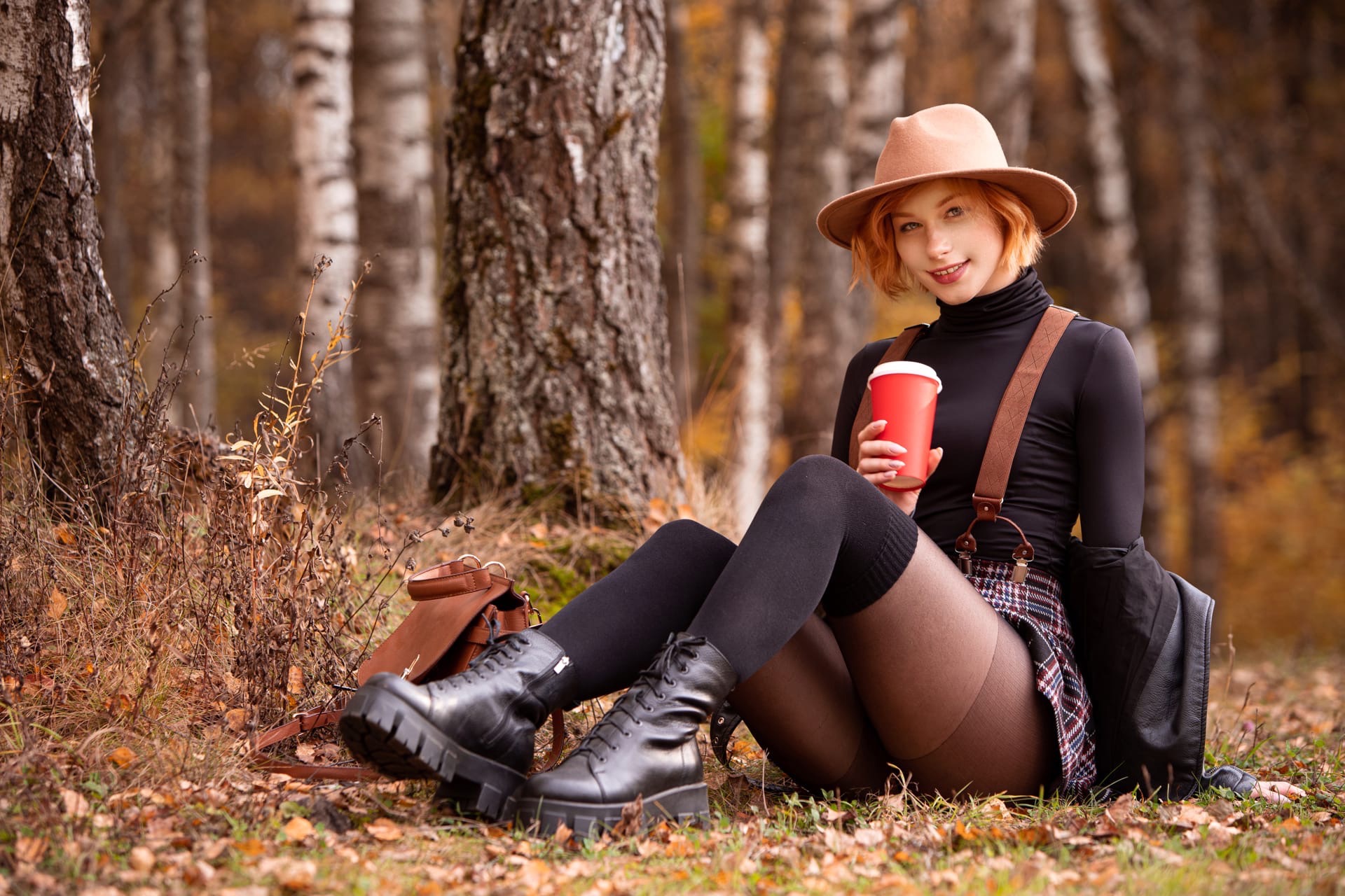 Модель Анастасия Жилина в шляпе, в приспущенной кожанке, в кофточке, в юбке и в чулках сидит на траве в осеннем лесу
