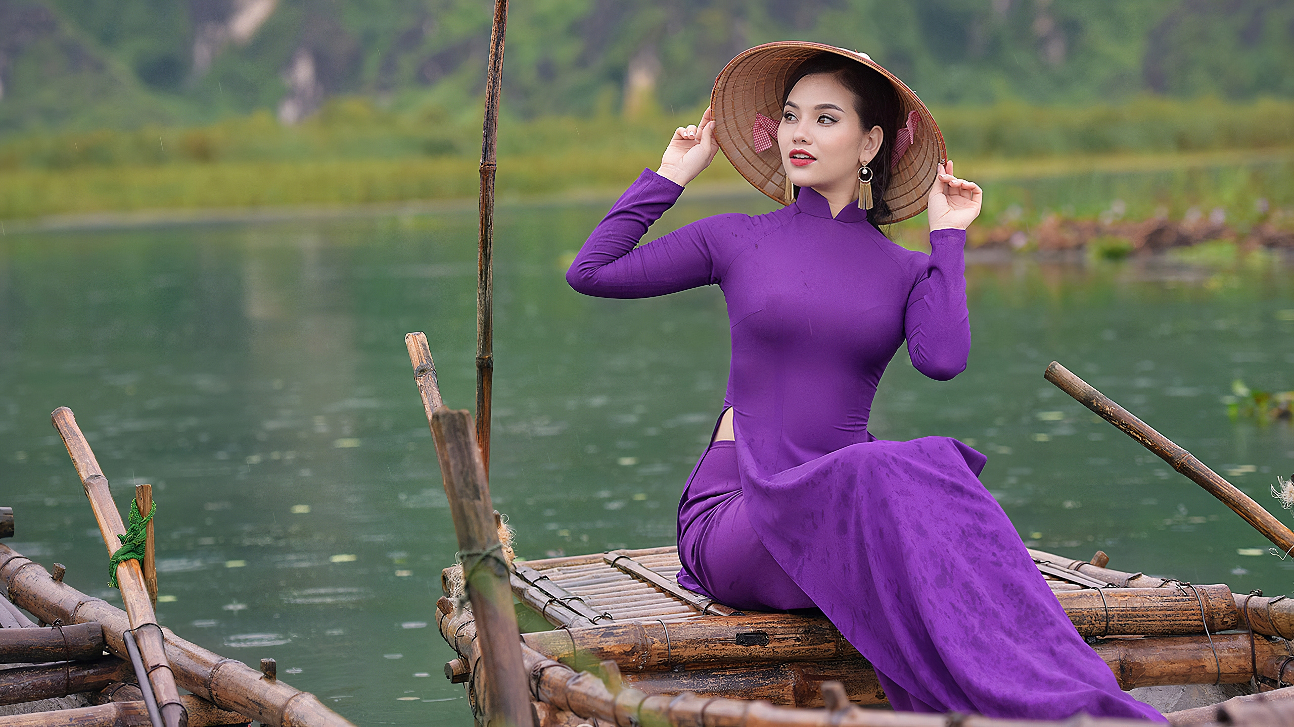 Девушка азиатка в национальной шляпе и в сиреневом платье сидит в лодке на фоне водоема и природы