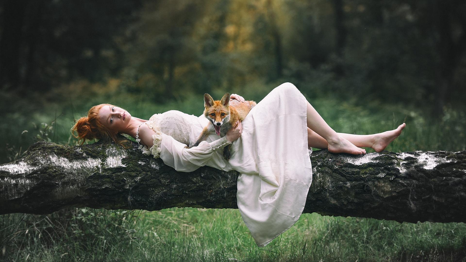 Рыжеволосая девушка в белом платье позирует, лежа на поваленном дереве с лежащей на животе лисой, на размытом фоне леса
