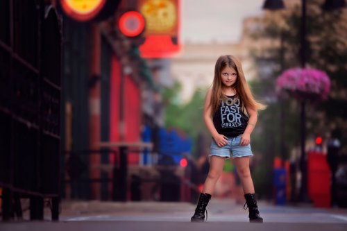 Девочка, в голубых шортах, черных майке и ботинках, стоит на улице города, расставив ноги