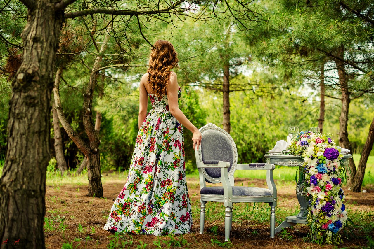Девушка с длинными вьющимися русыми волосами в летнем белом платье с яркими цветами стоит спиной к нам, положив руку на белое кресло