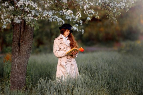 Рыжеволосая девушка в черной шляпе и в светлом плаще стоит на природе под цветущем деревом и читает держащую в реках книгу