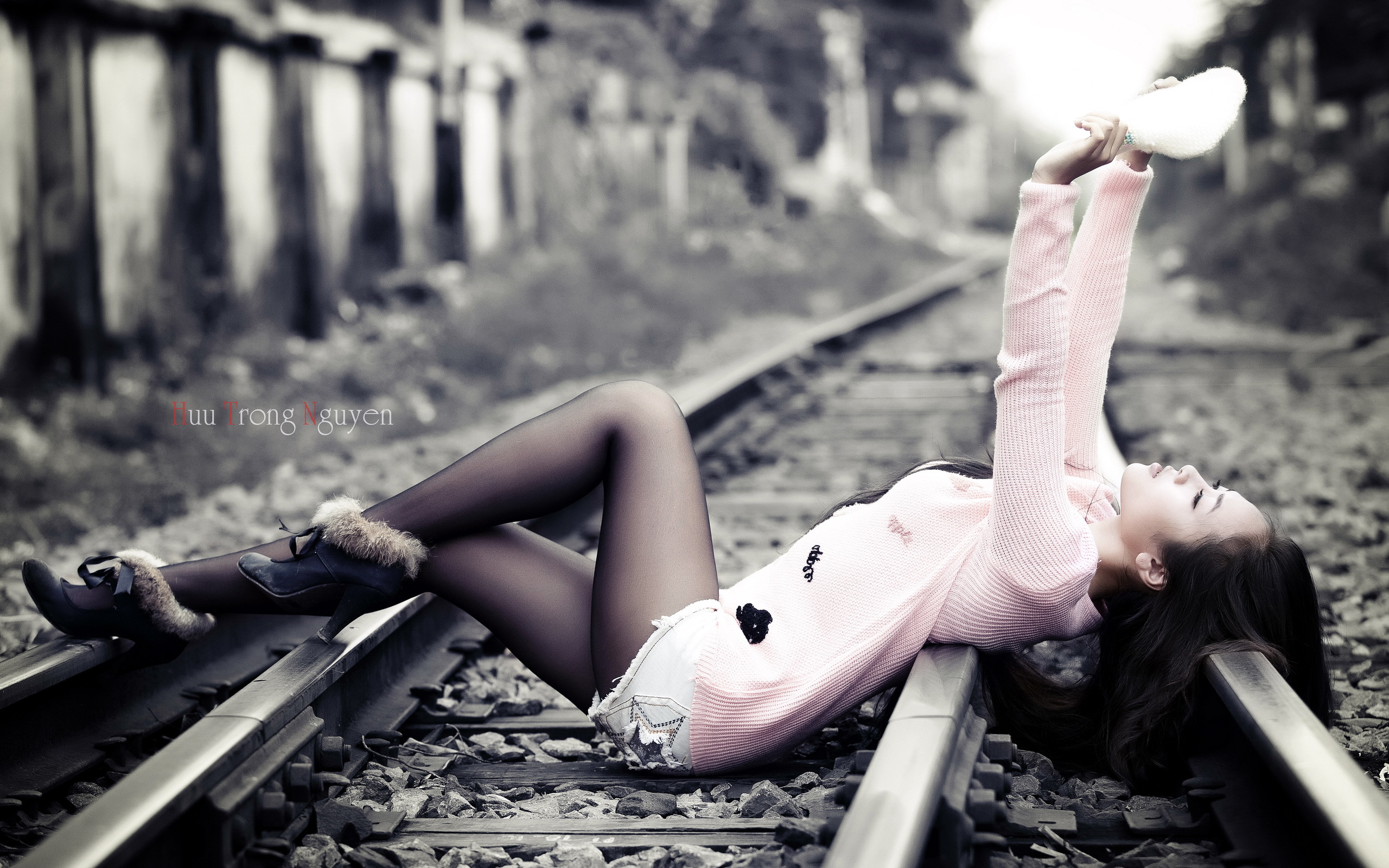 Девушка азиатка в нежно-розовом свитере, черных капроновых колготках и сапожках с мехом, лежит на рельсовых путях, подняв руки с чем-то белым