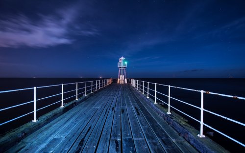 Светящий маяк, стоящий в конце деревянного пирса, огражденного металлическими перилами, расположенного на океанском побережье