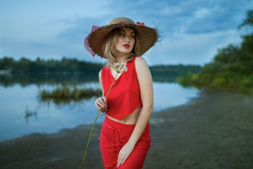Модель Nastya с цветком в руке, в шляпе, в красной блузке и в юбке стоит на берегу водоема