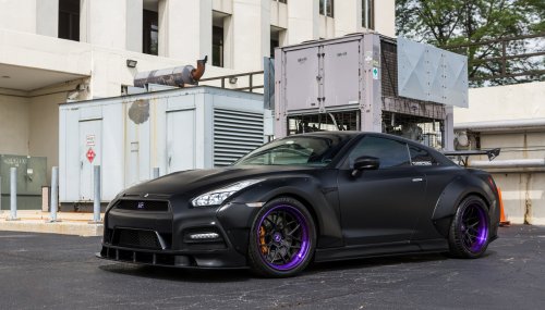 Черный матовый автомобиль Nissan GT-R с фиолетовыми дисками и оранжевыми тормозными суппортами припаркован на асфальтированной площадке