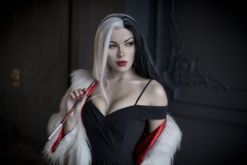 Девушка в образе Круэллы / Cruella из фильма Круэлла / Cruella позирует в черном платье и белой меховой накидке, держа мундштук, на фоне темной стены