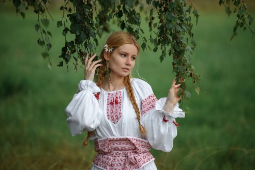 Модель Соня в белом с вышивкой сарафане стоит на природе и держится руками за ветки березы