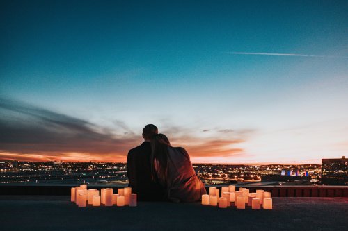 Мужчина и женщина сидят, обнявшись, на крыше, рядом горящие свечи, на фоне вечерней панорамы города с огнями и закатного неба