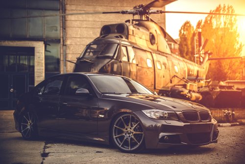 Темно-серый седан BMW E90 стоит на асфальте, перед большим старым военным вертолетом, на фоне здания и ярко освещенного закатным солнцем