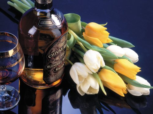 Бутылка виски Chivas Regal и бокал с напитком на переднем плане, рядом букет из желтых и белых тюльпанов на темном фоне