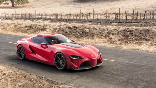Красный спортивный автомобиль Toyota Supra едет по асфальтированной дороге с желтой разметкой, на фоне сухих холмов с виноградником и редкими деревьям