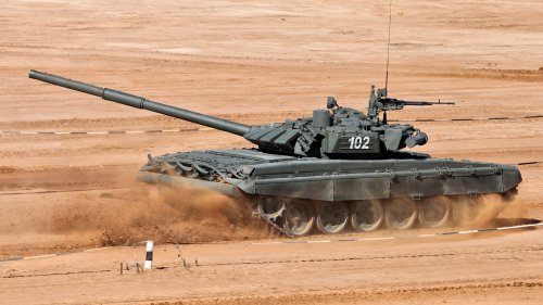Темно-зеленый танк с номером 102 на башне движется по песчаной местности, поднимая пыль вокруг гусениц, на фоне песочного ландшафта