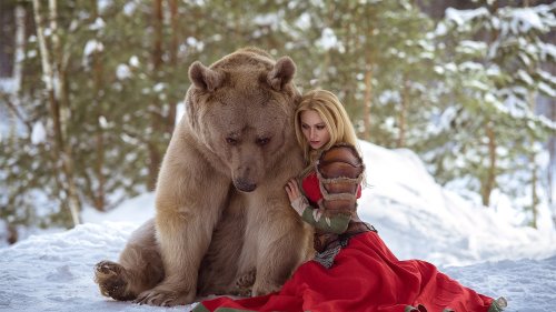 Блондинка в красном платье и коричневой куртке позирует прислонившись к бурому медведю