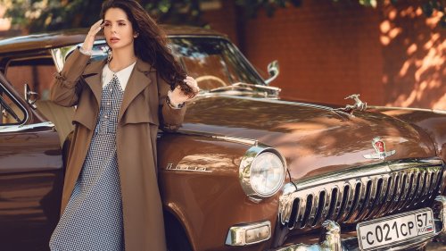 Девушка в коричневом плаще и клетчатом платье позирует рядом с коричневым автомобилем GAZ-21 Volga, Russiа на фоне кирпичной стены и листвы