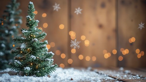 Маленькая елка, покрытая снегом с гирляндой, на заснеженной деревянной поверхности, на фоне размытой деревянной стены с белыми снежинками