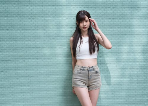 Молодая азиатская девушка в белом укороченном топе и светлых джинсовых шортах позирует, поправляя волосы рукой, на фоне мятной кирпичной стены