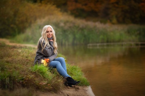 Блондинка в свитере и в джинсах сидит на берегу водоема и держит в руке пожелтевшие осенние листья